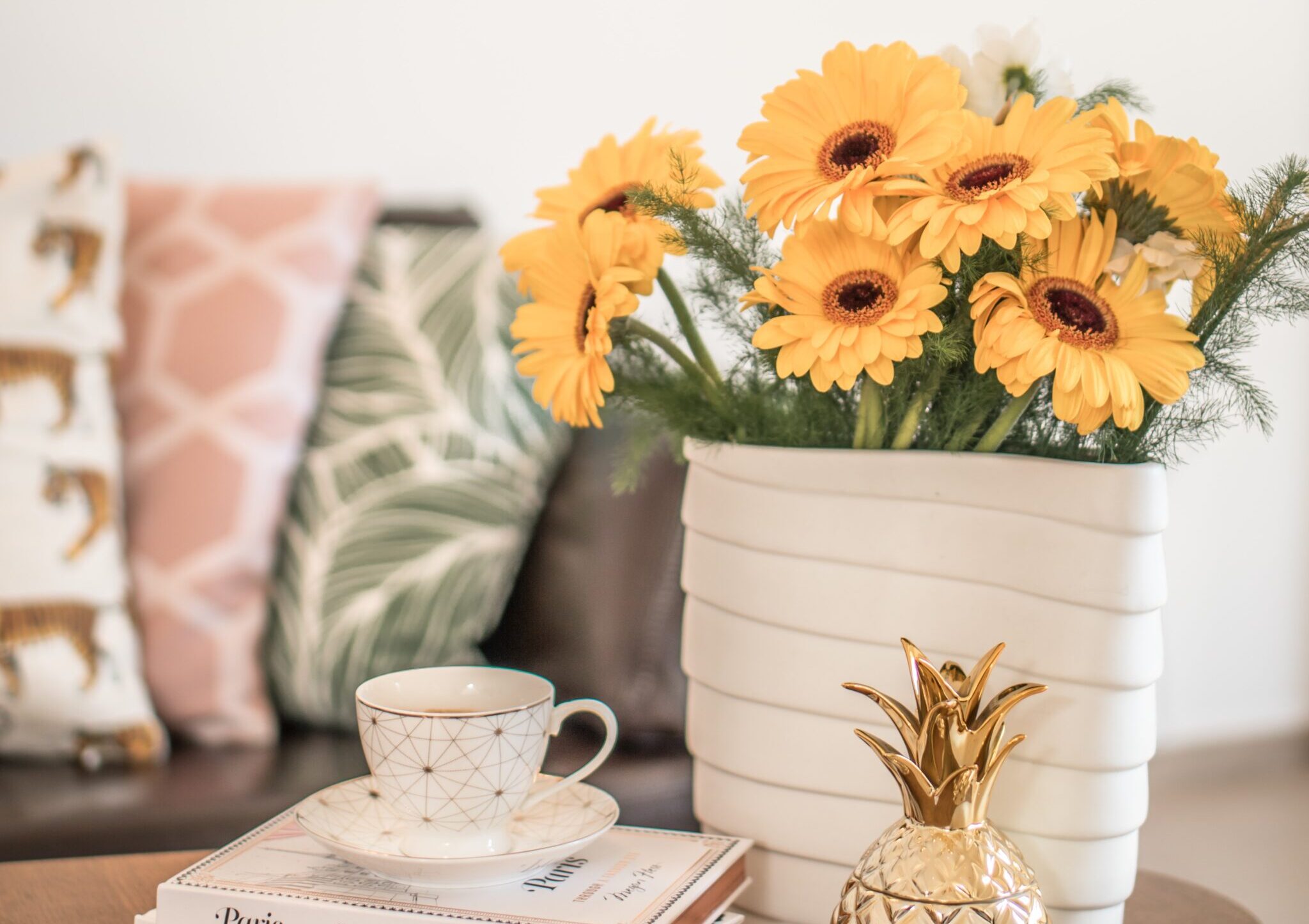 Bilden visar olika kuddar i en soffa tillsammans med gula blommor i en vas på ett bord. Blommiga kuddfodral till sovrum och vardagsrum hittar du online.